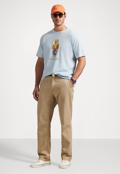 Gaiši zils kokvilnas T-krekls ar attēlu, kurā attēlots lācis žaketē, apvienots ar bēšiem kokvilnas biksēm un baltām sporta apaviem. Oranžs cepure un saulesbrilles.