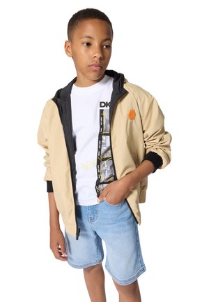 Veste légère beige avec accents noirs, fermeture éclair frontale et logo hexagonal. Portée sur un t-shirt graphique blanc et un short en jean.