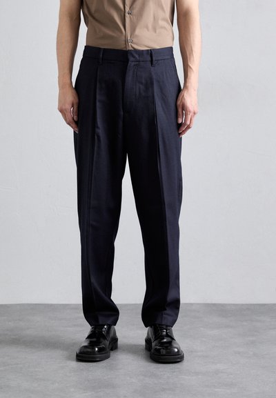 Navy trousers with a tailored fit, pleated front design, and belt loops. Worn with black shoes on a light gray surface.