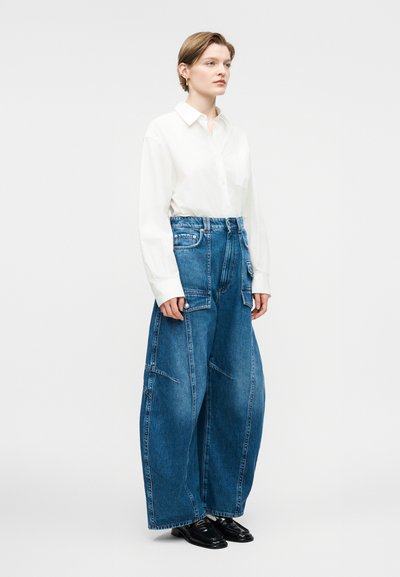 Άτομο που φοράει λευκό πουκάμισο με γιακά, μπλουζωμένο σε μπλε τζιν παντελόνι ψηλόμεσο και φαρδύ, με μπροστινές τσέπες, συνδυασμένο με μαύρα μοκασίνια.