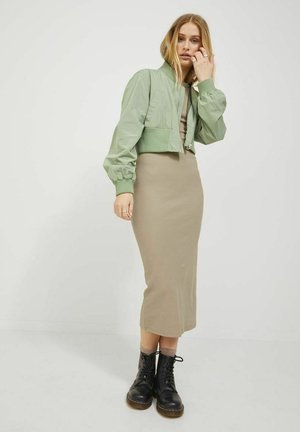 Chaqueta verde claro recortada con cremallera y puños elásticos, combinada con un vestido midi ajustado en tono beige y botas negras con cordones, sobre un fondo liso.