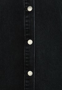 Tessuto di denim nero con una chiusura a bottoni verticale caratterizzata da bottoni a pressione argentati. Il tessuto presenta una texture liscia e un motivo di tessitura compatto.