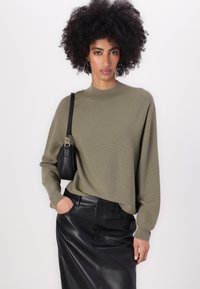 Olive green knit sweater with a high neckline and long sleeves, paired with a black leather skirt. Includes a small black bag on the shoulder.