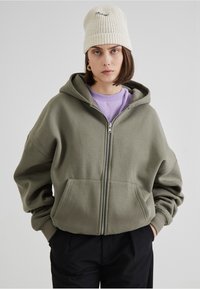 Sweat à capuche vert olive avec une fermeture éclair, poches avant et poignets côtes, superposé sur un t-shirt lavande, porté avec un bonnet en tricot blanc.