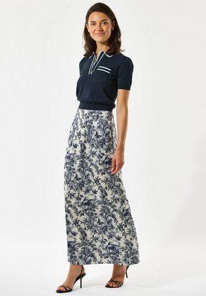 Žena oblečená v námornícky modrom tričku s krátkym rukávom a bielej maxi sukni s modrými botanickými vzormi, stojaca v čiernych lodičkách.