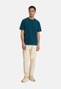 Törökzöld, rövid ujjas póló, kerek nyakkivágással, bézs nadrággal és világos sportcipővel párosítva. Sima anyag, laza szabás, minimalista design.