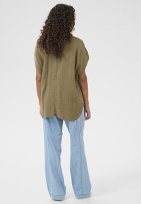 Olive green, short-sleeve blouse with a textured finish. Worn with light blue wide-leg jeans, featuring side seams and a relaxed fit.