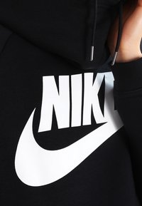Schwarzes Sweatshirt mit einem großen weißen Nike-Logo. Mit Kapuze und Kordeln ausgestattet, aus einem weichen, strukturierten Stoff. Nahaufnahme.