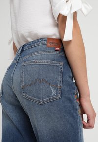 Siniset farkut korkealla vyötäröllä, joissa ommellut takataskut ja nahkainen merkintä. Kangas näyttää kuluneelta ja koristeelliselta kirjonnalta.