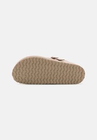 Beige mocka sko med en strukturerad gummisula med ett zigzag-mönster för grepp. Rundad form med en rosett detalj på toppen.