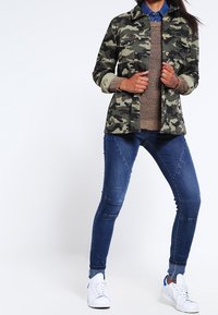 Chaqueta de estampado camuflaje con cuello, suéter de punto beige y jeans skinny de mezclilla azul, combinados con zapatillas blancas.