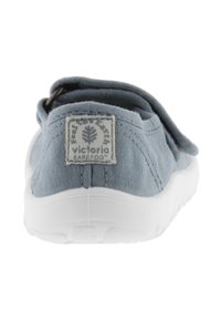 Zapatilla de lona azul claro tipo slip-on con suela de goma blanca, que presenta un parche con el logo en el talón y un lazo para facilitar su colocación.