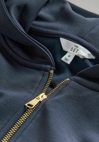 Felpa blu navy con una cerniera dorata, caratterizzata da una trama morbida e un'etichetta con scritto "THE SET M" all'interno del colletto.