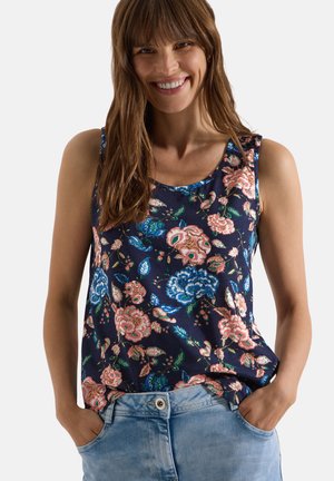 Femme portant un haut sans manches bleu marine avec un imprimé floral rose et bleu et un jean bleu clair, souriante avec les mains dans les poches.