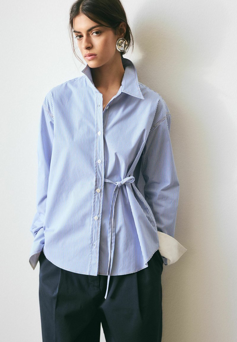 Žena v nadrozmernej modro-bielej pruhovanej košeli so zaviazaným detailom na boku a čiernych nohaviciach stojaca oproti svetlej stene.