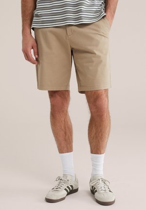 Férfi bézs rövidnadrágban, fehér zokniban, bézs és fekete tornacipőben, csíkos ingben, kezek a zsebekben.