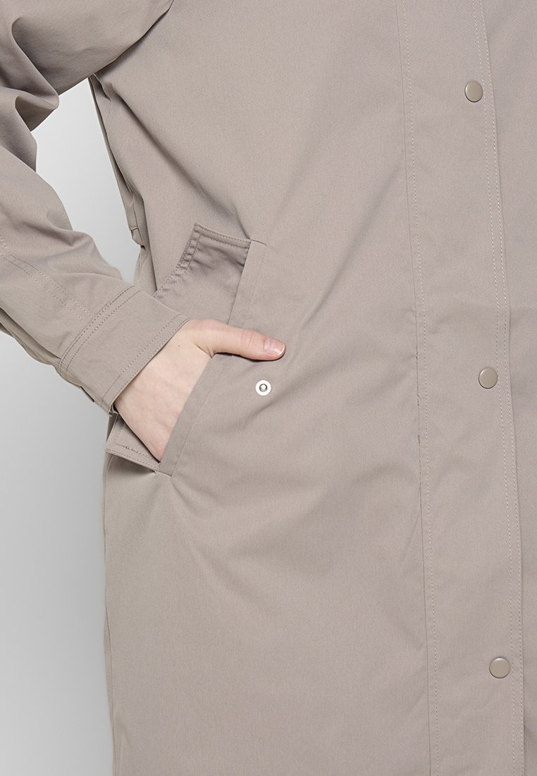 Personne portant un manteau beige avec une main dans la poche avant, boutons-pression en métal visibles et coutures sur les poignets et la poche.