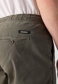Pantaloni khaki Ecoalf cu talie elastică, buzunar spate cu buton și o etichetă neagră. Textură de țesătură moale și netedă.