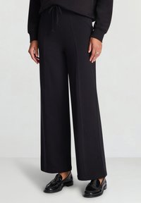 Schwarze Hosen mit weitem Bein und weicher Textur, ausgestattet mit einem elastischen Bund und Kordelzug. Kombiniert mit glänzenden schwarzen Loafers.