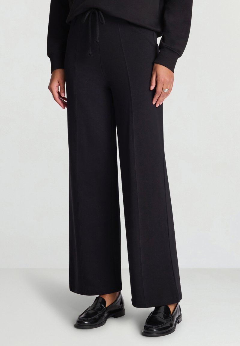 Schwarze Hosen mit weitem Bein und weicher Textur, ausgestattet mit einem elastischen Bund und Kordelzug. Kombiniert mit glänzenden schwarzen Loafers.