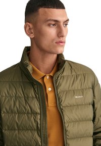 Chaqueta acolchada verde con cuello alto, cierre de cremallera y logotipo blanco. Lleva puesta una camiseta polo mostaza con tres botones.