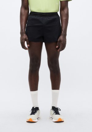 Juodi sportiniai šortai su elastiniu liemeniu, derinami su baltais kojinėmis ir šviesiais bėgimo batais su oranžiniais akcentais.