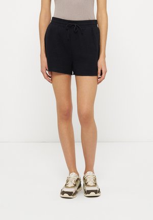 Naine kannab musta tekstuuriga nööriga lühikeseid pükse, beeži sõrmikut ja valgeid ning pruune tossusid, seistes ühevärvilise valge tausta ees.