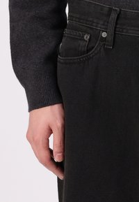 Svart denimjeans med smal passform, med lomme og metallknappedetaljer. Hånden hviler på sidesømmen. Genser i mørkegrå.