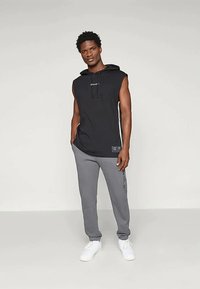 Black sleeveless hoodie with a front logo and gray sweatpants featuring side lettering, paired with white sneakers on a neutral background.