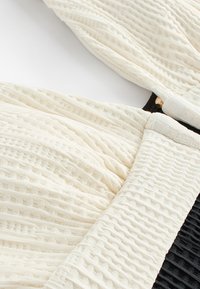 Detailný záber bielej textúrovanej látky s vlnitými vzormi a zoskupenými detailmi na čiernom textúrovanom pozadí.