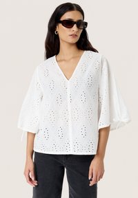 Hvid bluse med diamantudskæringsmønster, V-hals, ballonsleeves og bånddetaljer ved manchetterne. Sammen med mørke jeans.