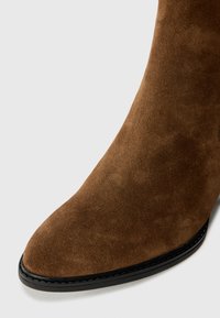 Gros plan d'une botte en daim marron avec un bout pointu et une semelle noire cousue sur un fond gris clair.