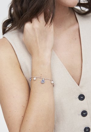 Vrouw draagt een beige mouwloos topje met knopen, toont haar arm met een zilveren armband met parels en blauwe bloembedels.