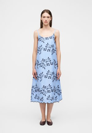 Mujer con un vestido midi azul claro con tirantes finos y bordado floral oscuro, combinada con zapatos planos marrón oscuro, de pie sobre un fondo blanco.