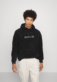 Hanorac negru din fleece cu o suprafață texturată, glugă cu șnur și buzunar frontal tip kangaroo, având inscripții albe în japoneză. Portat cu pantaloni deschiși.