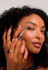 Make-upkwast met een zwart handvat dicht bij het oog gehouden, terwijl product wordt aangebracht. Model heeft krullend haar, gouden ringen en een gladde, stralende huid.