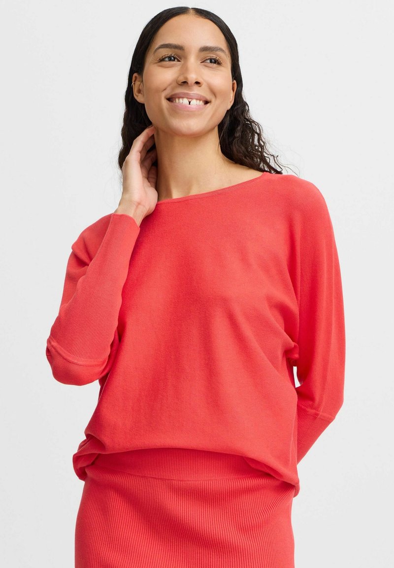 Pull en tricot corail à manches longues, avec une coupe ample et un col bateau. Texture douce et poignets côtelés. Porté avec une jupe côtelée assortie.