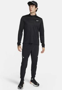 Crni sportski outfit koji se sastoji od dukserice s dugim rukavima koja se zatvara na zip, i usklađenih hlača, oboje s bijelim Nike logotipom, upotpunjen crnim tenisicama.