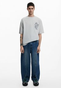 Világosszürke, oversized t-shirt, sötét hímzett logóval a bal oldalon. Széles lábú kék farmernadrággal és zöld részletekkel ellátott fekete sportcipővel párosítva.