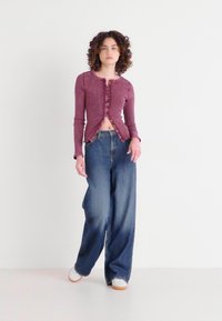 Long-sleeve maroon cropped top with textured fabric, button front, paired with wide-leg blue jeans and white sneakers on a plain background.