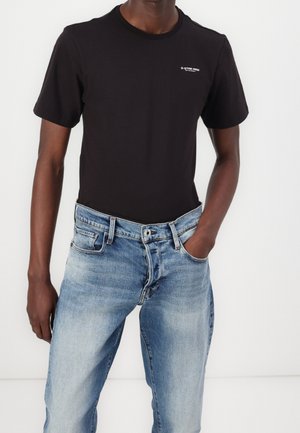 Homme portant un t-shirt noir uni à manches courtes avec un petit logo sur la poitrine et un jean bleu clair délavé, une main dans la poche.