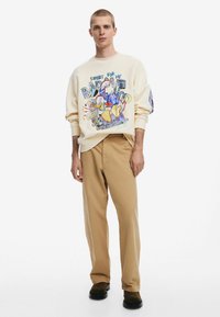 Crème sweatshirt met grafische print en tekst, gecombineerd met beige broeken. Model draagt zwarte schoenen; de stof lijkt zacht met een ontspannen pasvorm.