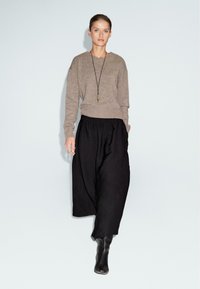Maglione lavorato a maglia di colore marrone chiaro con una vestibilità rilassata, abbinato a pantaloni neri a gamba larga e fluida. Accessoriato con una lunga collana con pendente.