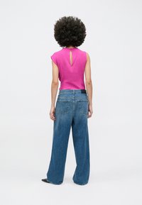 Persoană cu păr creț purtând un top roz fără mâneci și blugi albaștri cu croială largă, stând cu spatele spre privitor pe un fundal alb.