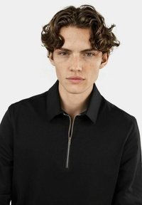 Czarny polo z zamkiem błyskawicznym wykonany z miękkiego materiału z kołnierzykiem. Posiada srebrny zamek i czysty, minimalistyczny design. Brak widocznych wzorów lub akcentów.