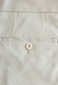 Tissu beige avec une poche horizontale fendue cousue et un seul bouton assorti à quatre trous cousu sous l'ouverture de la poche.