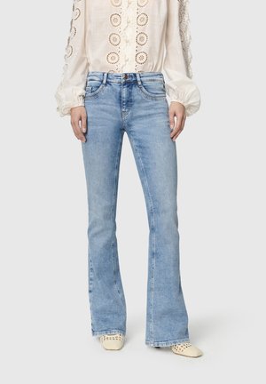 Vrouw draagt lichtblauwe wijde jeans gecombineerd met een wit blouse met lange mouwen met kanten uitsnijdingen en crèmekleurige geweven puntige schoenen.