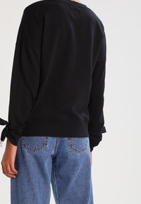 Sudadera negra con mangas largas y lazos de cinta en los puños, combinada con jeans de tiro alto en azul claro. Textura suave y corte holgado.