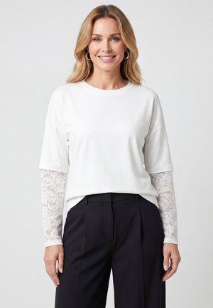 Uśmiechnięta kobieta o blond włosach nosi biały top z koronkowymi długimi rękawami pod spodem oraz czarne spodnie z wysokim stanem na jednolitym tle.