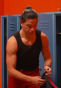 Schwarzes Tanktop und rote Shorts getragen von einer muskulösen Person, die neben blauen Schließfächern in einer orangefarbenen Umgebung steht und ihre Kleidung richtet.